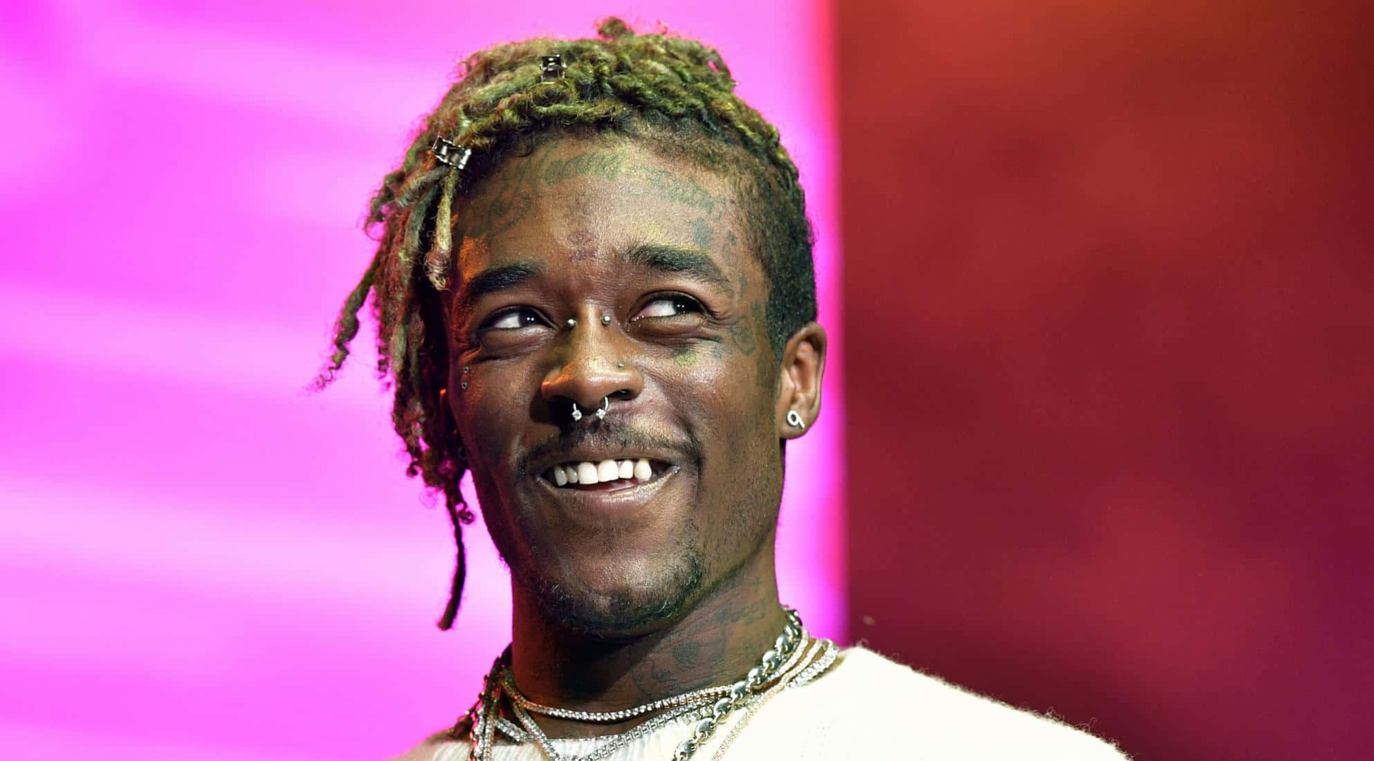 Lil Uzi Vert 桌布 壁紙 — 一個微笑的男人，留著綠色辮子，有多個穿孔，背景是充滿活力的粉紅色和紅色漸變。