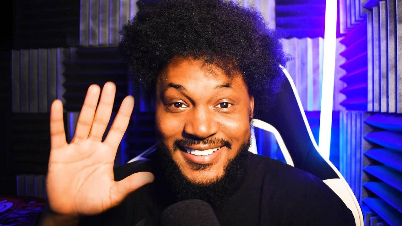 Coryxkenshin 桌布 壁紙 — 一個微笑的男人，留著非洲式髮型，在藍色燈光的遊戲設置中，隔音牆壁，愉快地揮手。
