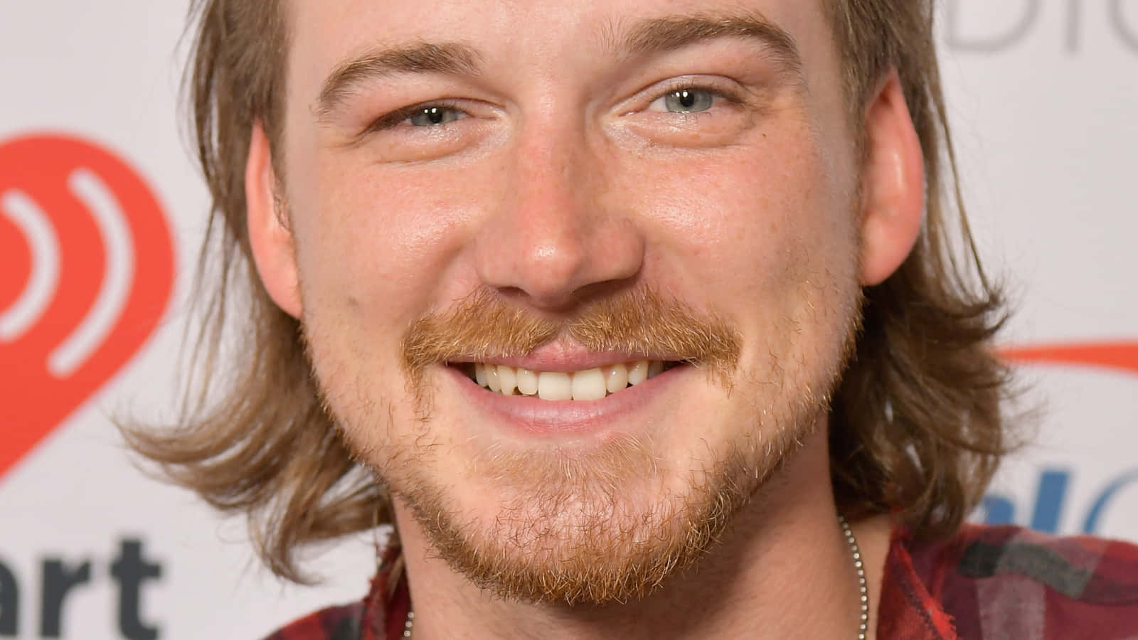 Morgan Wallen 桌布 壁紙 — 一個微笑的男人，留著鬍子和及肩長髮，在品牌背景前的特寫肖像。