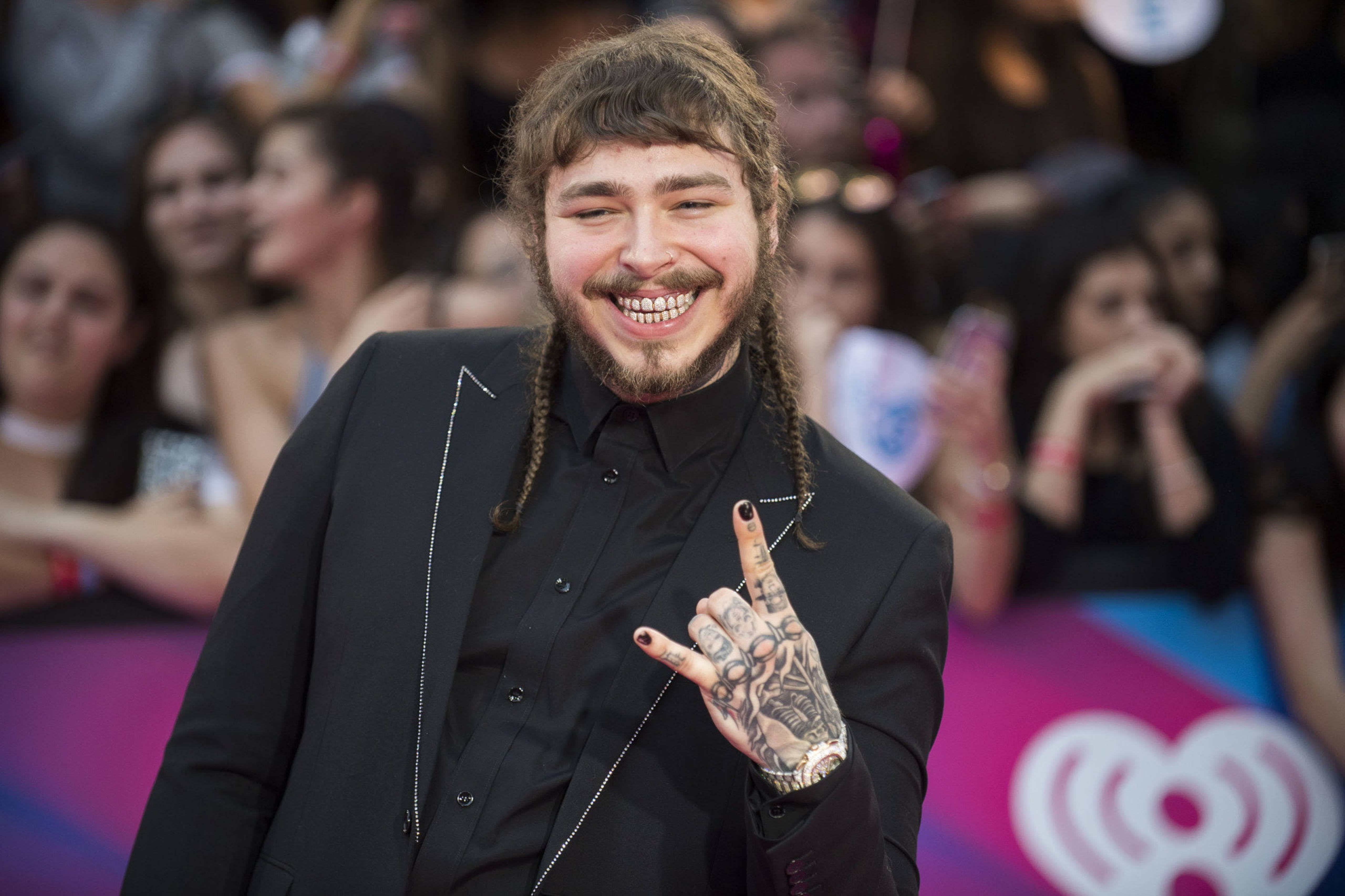 Post Malone 2017 iHeartRadio Awards 壁紙 — 一個微笑的男人，編著辮子，有紋身，在明亮的燈光下擁擠的活動中比出和平手勢。