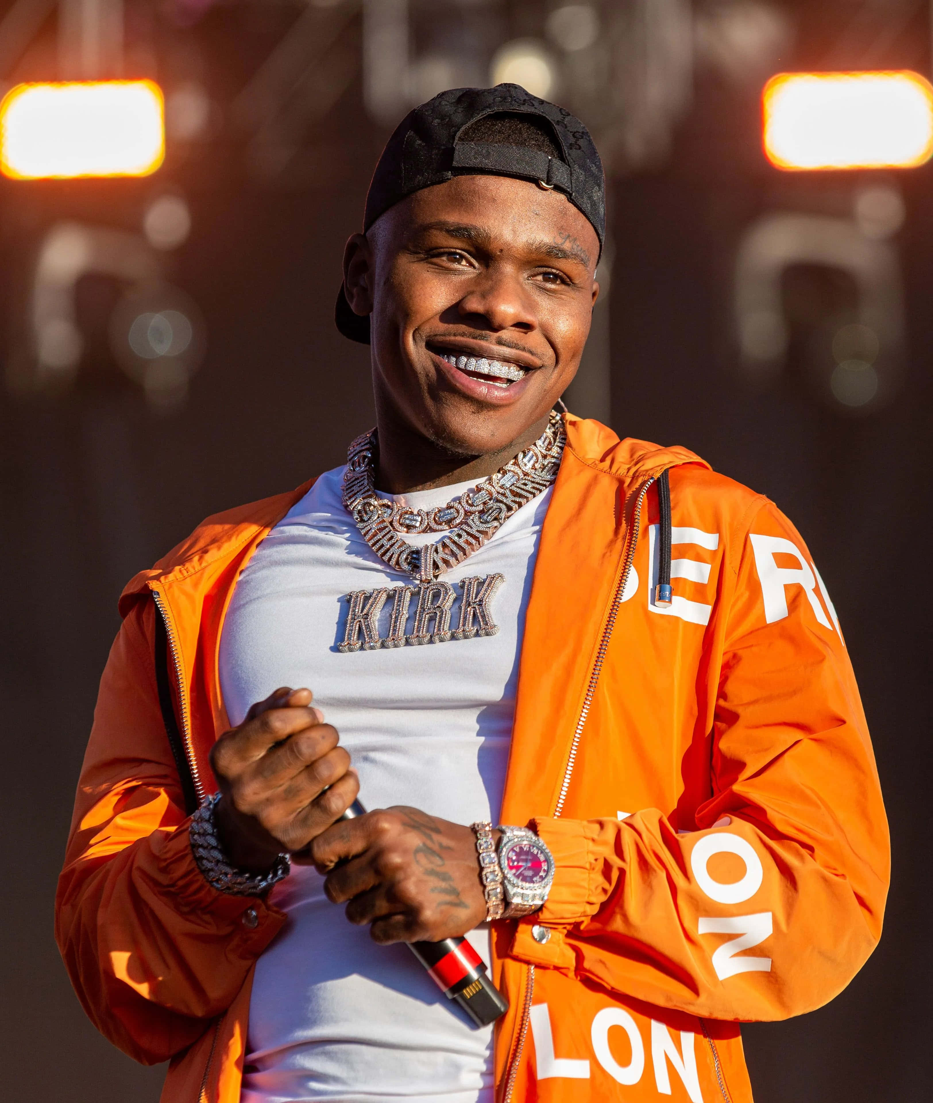 Dababy 桌布 壁紙 — 一個微笑的男人，身穿橙色外套和黑色帽子，在舞台燈光下拿著麥克風。