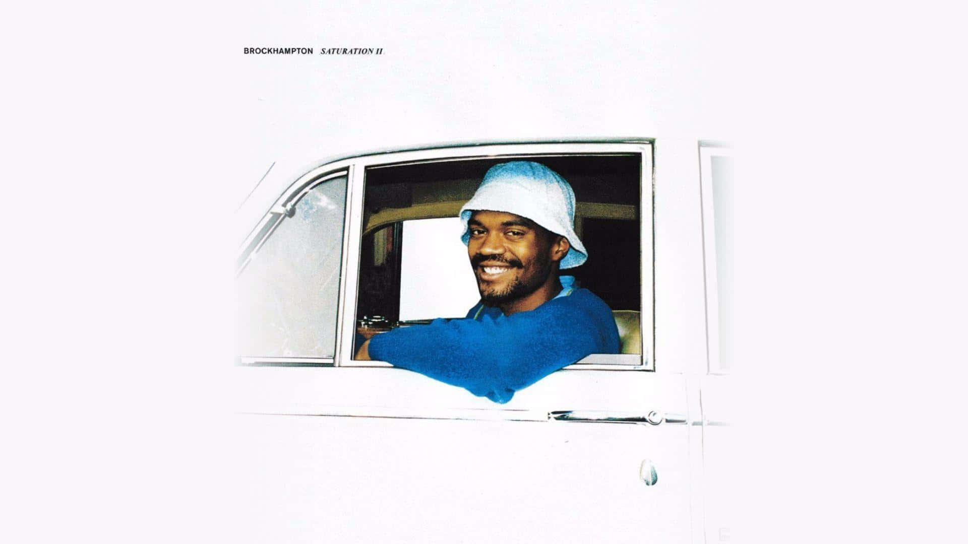 Brockhampton 桌布 壁紙 — 一個微笑的男人，身穿藍色毛衣和漁夫帽，從白色汽車的車窗探出頭來，背景是純白色。