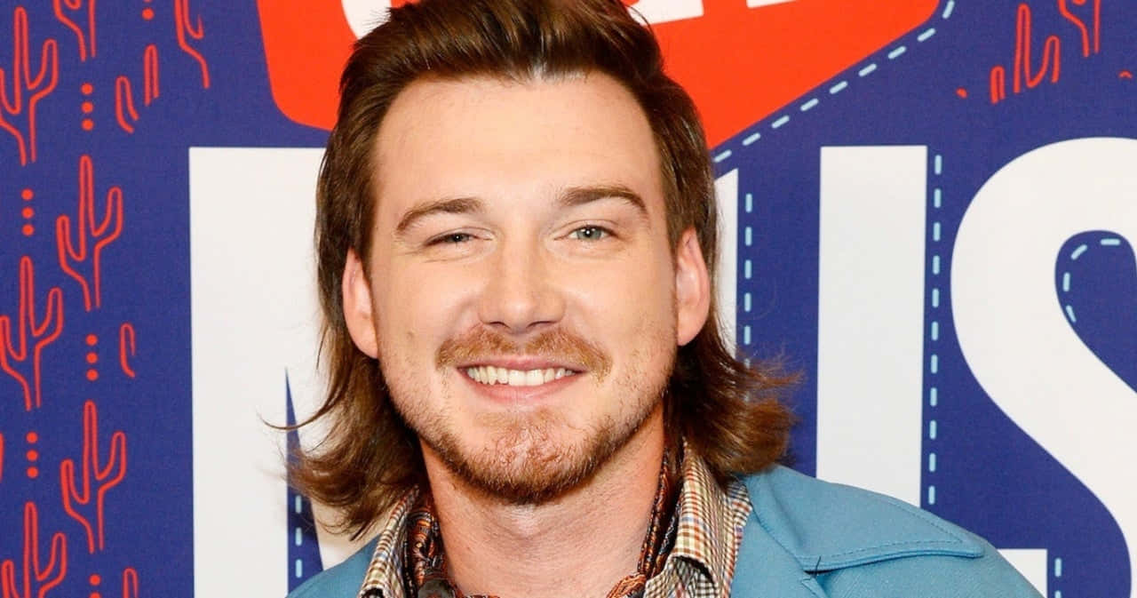 Morgan Wallen 桌布 壁紙 — 一個微笑的男人，長長的棕色頭髮和鬍鬚，穿著藍色外套，背景是色彩繽紛的仙人掌圖案。