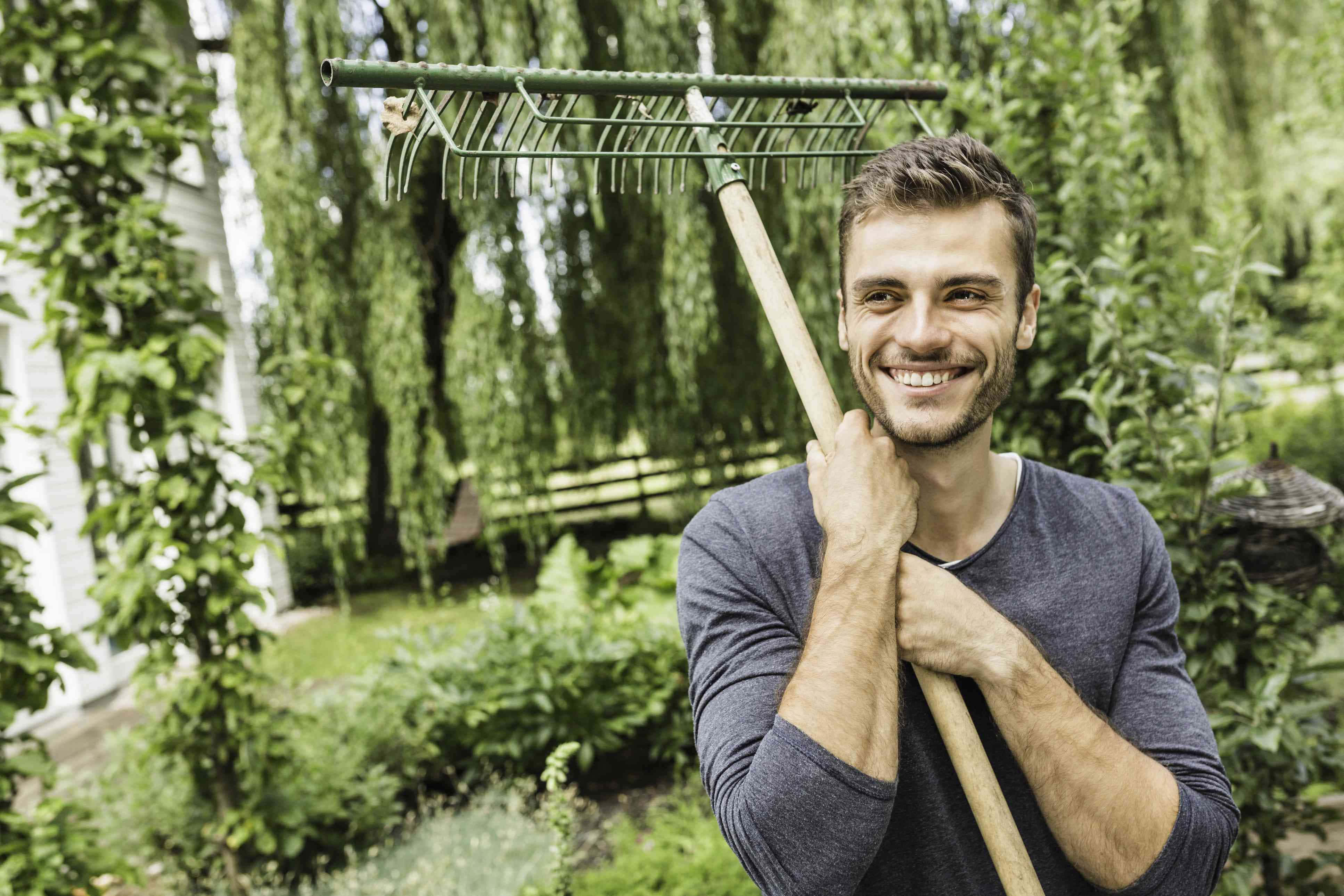 Gardening 桌布 壁紙 — 一個微笑的男人在一個鬱鬱蔥蔥的綠色花園裡拿著一把綠色耙子，背景有垂柳和一間白色房子。