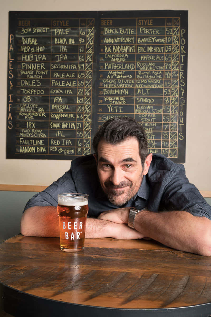 Ty Burrell 桌布 壁紙 — 一個微笑的男人將手臂擱在木桌上，面前放著一杯啤酒，身後是黑板啤酒菜單，身處舒適的酒吧。
