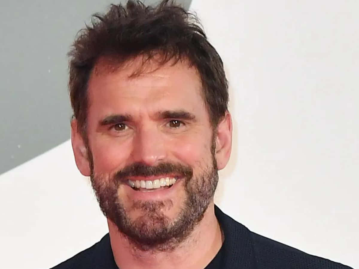 matt dillon 桌布 壁紙 — 一個微笑的男人特寫肖像，深色頭髮和鬍鬚，穿著深色襯衫，背景簡單。