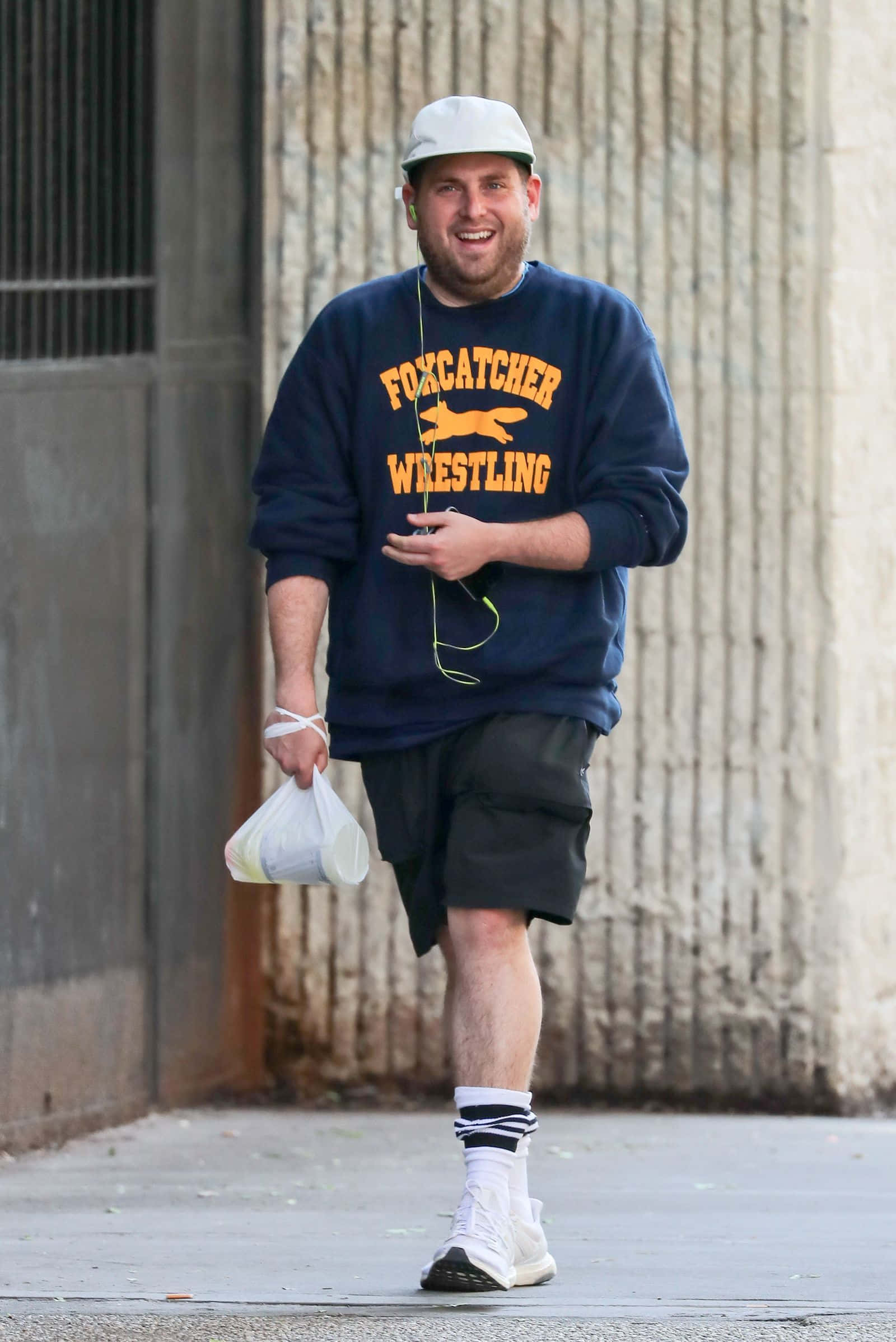 jonah hill 桌布 壁紙 — 一個微笑的男人穿著海軍藍色 'Foxcatcher Wrestling' 運動衫和白色帽子，拿著一個膠袋走在混凝土小巷中。