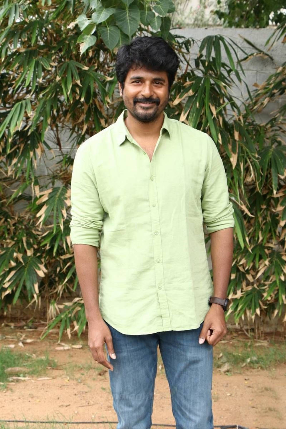 Sivakarthikeyan 桌布 壁紙 — 一個微笑的男人穿著淺綠色襯衫和牛仔褲，自信地站在茂密的綠色竹葉背景下。