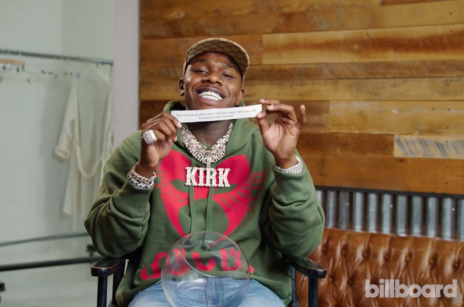 Dababy 桌布 壁紙 — 一個微笑的男人穿著綠色連帽衫，上面有紅色心形和 'KIRK'，坐在椅子上，背後是木牆，手持一張白色卡片。