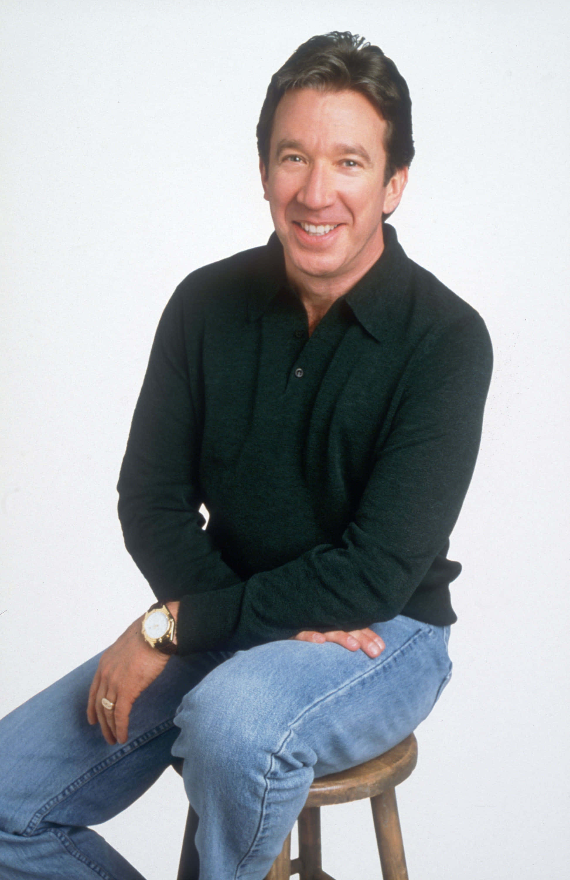 Tim Allen 桌布 壁紙 — 一個微笑的男人穿著黑色Polo衫和藍色牛仔褲，隨意地坐在白色背景前的木凳上。