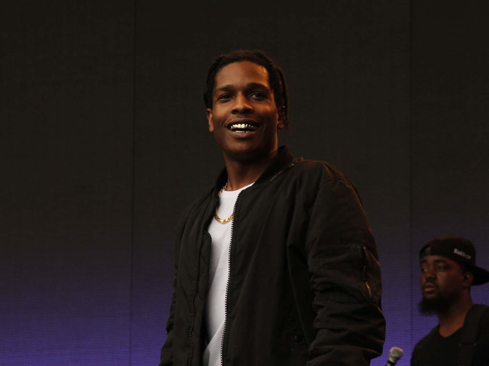 ASAP Rocky 壁紙 — 一個微笑的男人穿著黑色外套和白色T恤，站在一個背後有紫色燈光的舞台上。