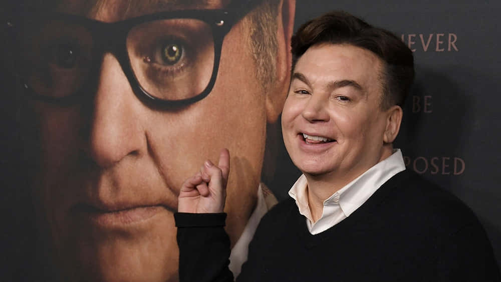 Mike Myers 桌布 壁紙 — 一個微笑的男人穿著黑色毛衣，站在一張戴著眼鏡的自己臉部大 poster 前。