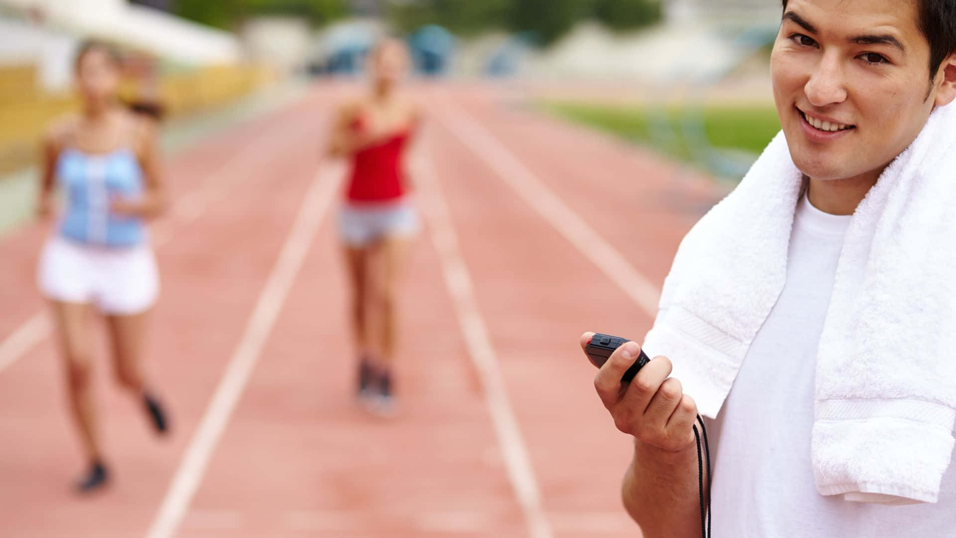 Best Sports 桌布 壁紙 — 一個微笑的男人脖子上掛著毛巾，在紅色跑道上拿著計時器，身後有模糊的跑者。