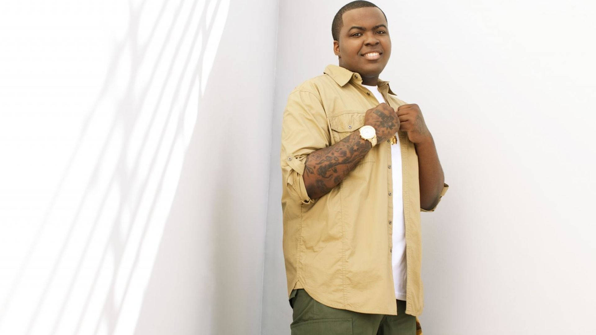 Sean Kingston In Photoshoot 壁紙 — 一個微笑的紋身男子在帶有對角光線圖案的白色牆壁前調整他的米色襯衫。