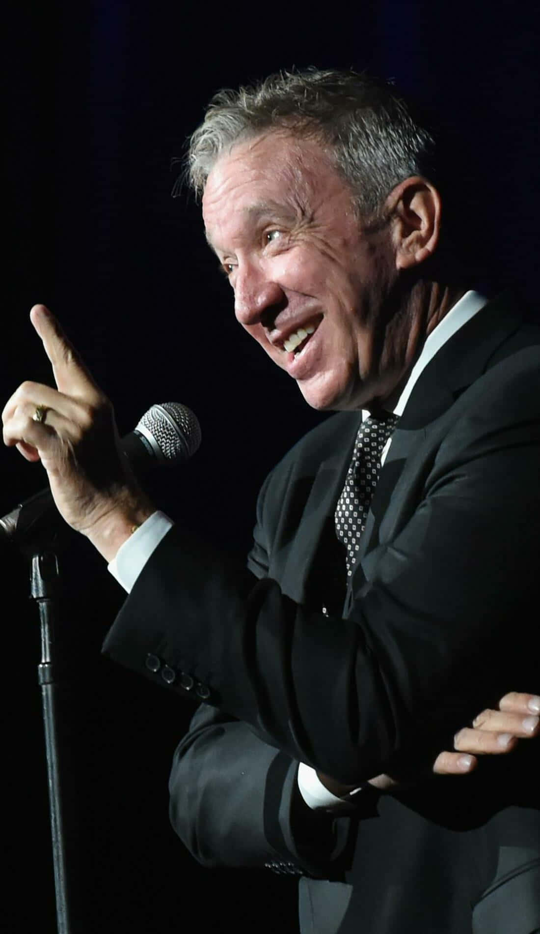 Tim Allen 桌布 壁紙 — 一個微笑的西裝男子在黑暗的舞台上熱情地指著，同時拿著麥克風。