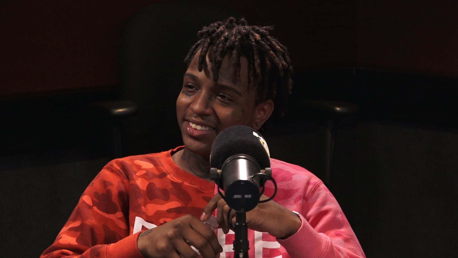 trippie redd 桌布 壁紙 — 一個微笑的辮子頭人物穿著紅色迷彩運動衫，對著錄音室麥克風說話。