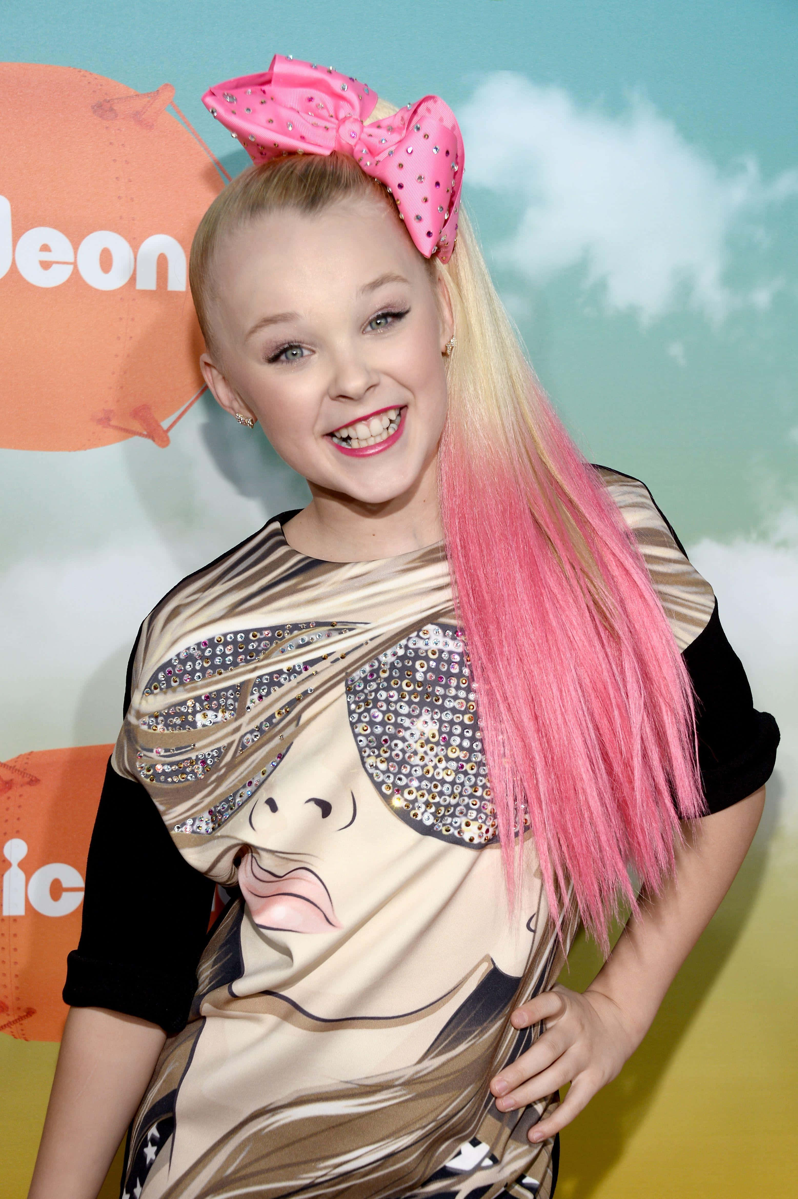 jojo siwa 桌布 壁紙 — 一個微笑的金髮女孩，戴著巨大的粉紅色蝴蝶結，穿著帶有卡通臉設計的閃亮服裝。