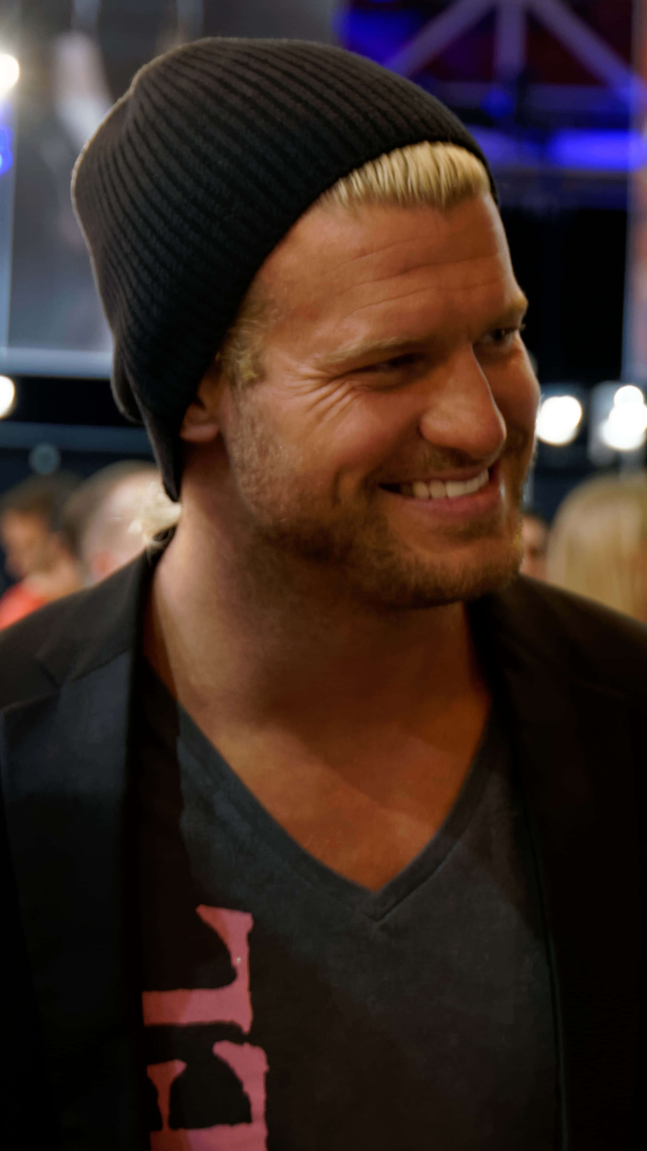 Dolph Ziggler Stage Stand-up Comedian 壁紙 — 一個微笑的金髮男子戴著黑色無邊帽，在昏暗的活動空間中，身後有模糊的燈光，溫暖地笑著。