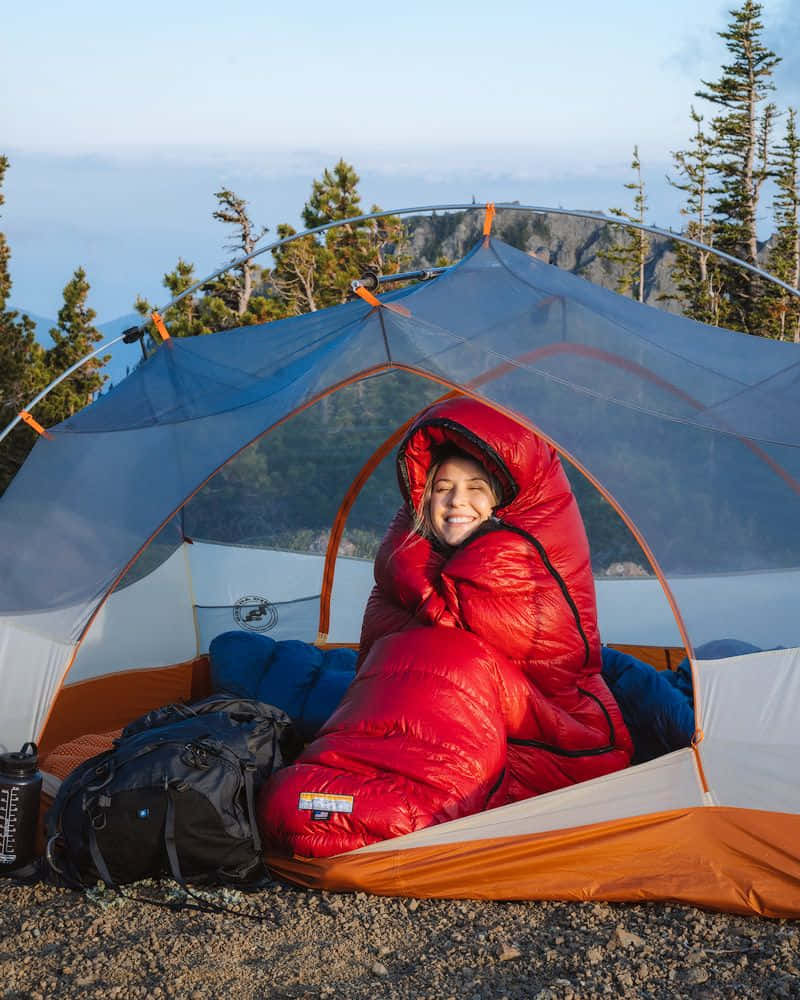 Winter Hiking 桌布 壁紙 — 一個微笑的露營者躺在紅色睡袋裡，舒適地坐在藍色帳篷中，欣賞日出時的山景。