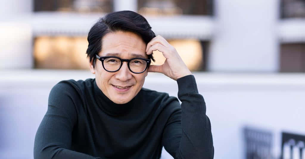 Ke Huy Quan 桌布 壁紙 — 一個微笑的黑髮戴眼鏡男士，將手放在頭上，背景是柔和模糊的現代建築。