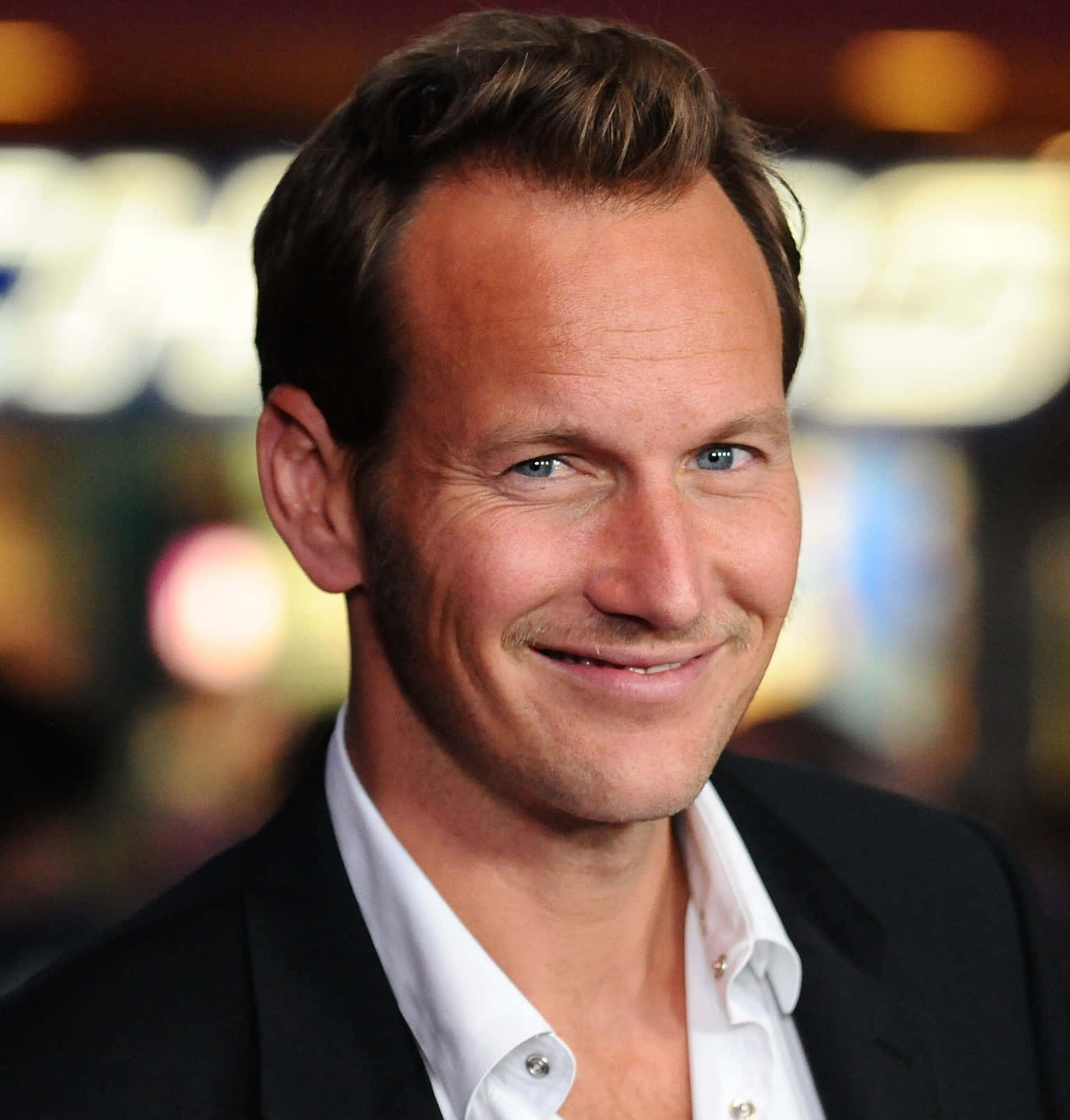 Patrick Wilson 桌布 壁紙 — 一個微笑藍眼男士的近距離肖像，身穿黑色西裝和白色襯衫，背景是模糊的溫暖燈光。