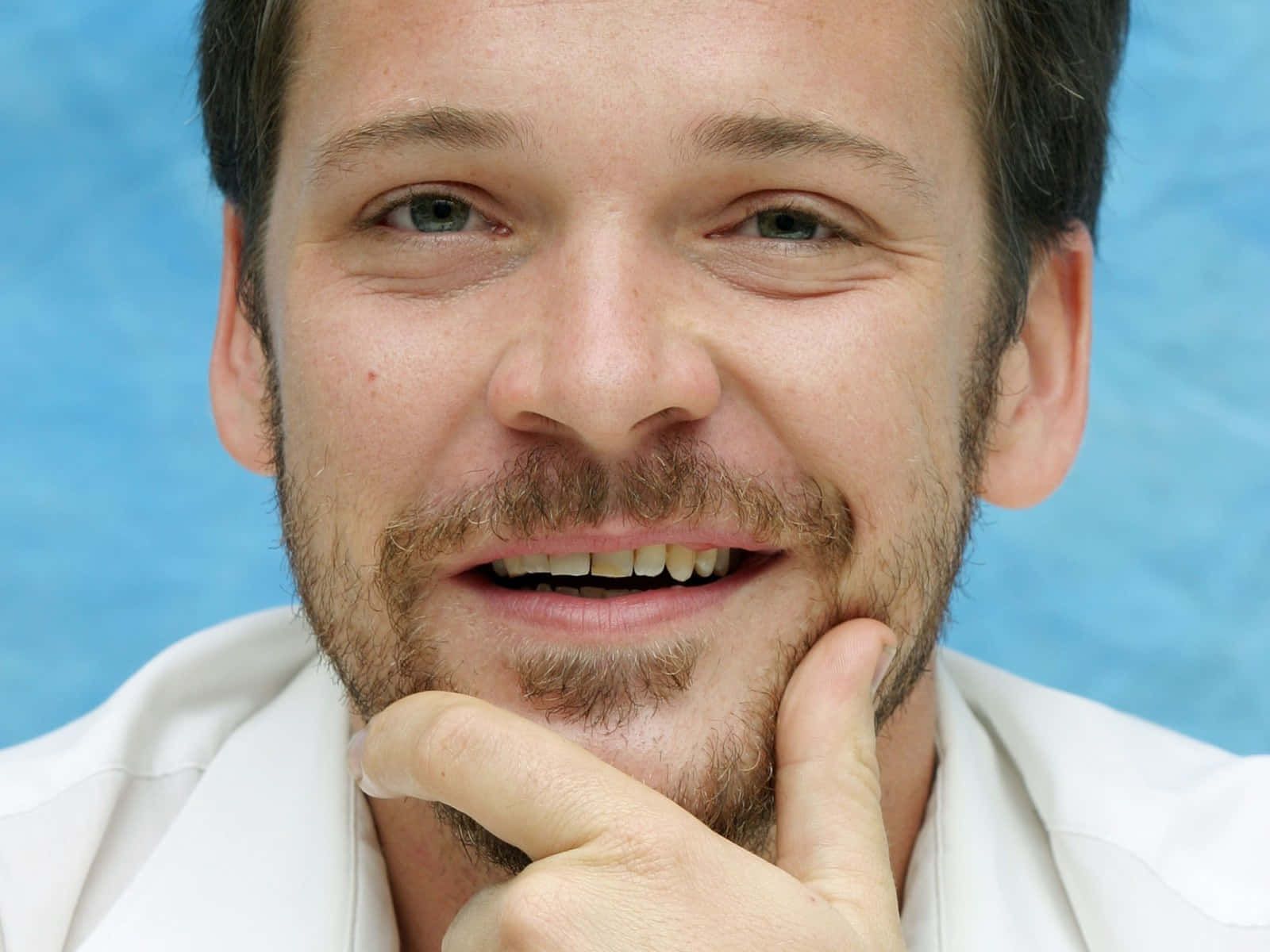 Peter Sarsgaard 桌布 壁紙 — 一個微笑鬍鬚男嘅特寫肖像，手放喺下巴，背景係藍色。