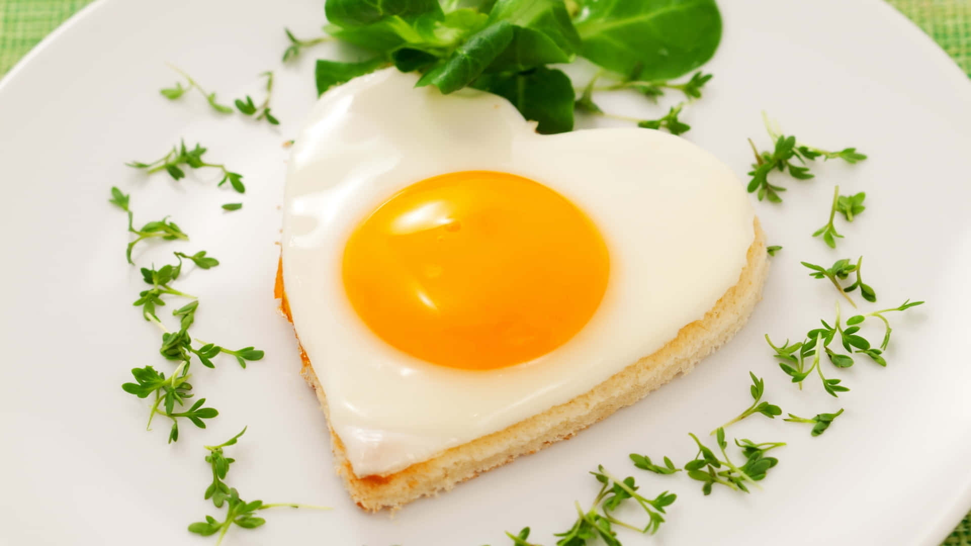 Egg White 桌布 壁紙 — 一個心形太陽蛋，蛋黃鮮豔，放在多士上，配以新鮮綠色香草裝飾，置於白色碟上。