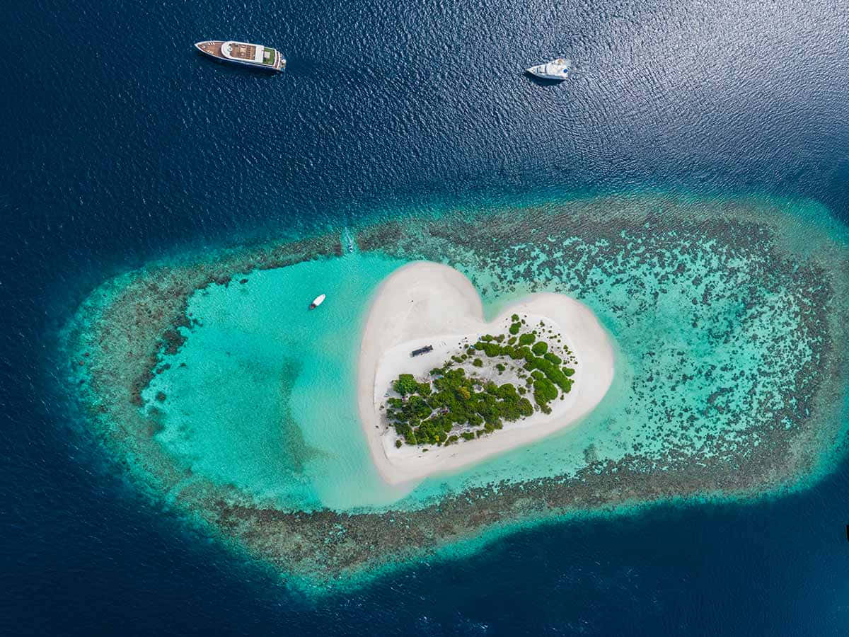 Maldives Island 桌布 壁紙 — 一個心形熱帶島嶼的鳥瞰圖，擁有白色沙灘和綠色樹木，周圍環繞著碧綠的潟湖和深藍色的海洋，附近有船隻。