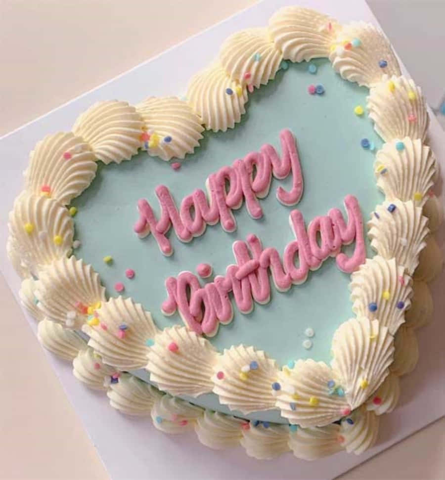 生日蛋糕 桌布 壁紙 — 一個心形生日蛋糕，帶有淺藍色糖霜和粉紅色擠花文字，周圍環繞著白色貝殼邊緣和彩色糖粒。