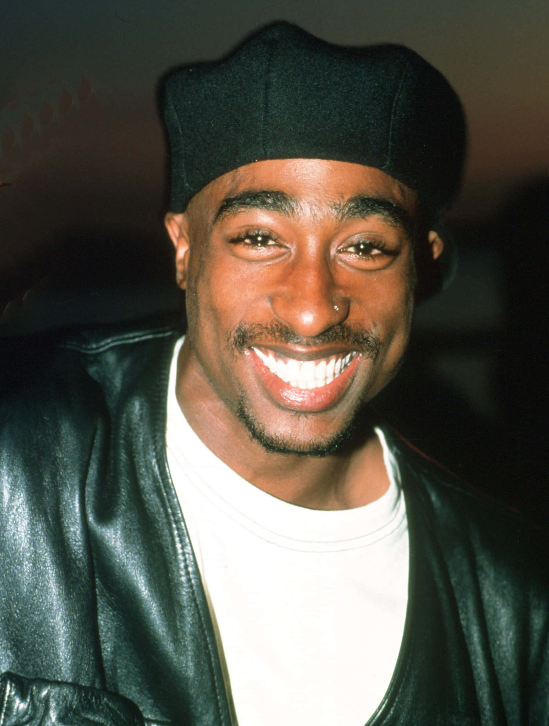Tupac 桌布 壁紙 — 一個快樂嘅男人，咧嘴大笑，戴住黑色冷帽同皮褸，背景係深色。
