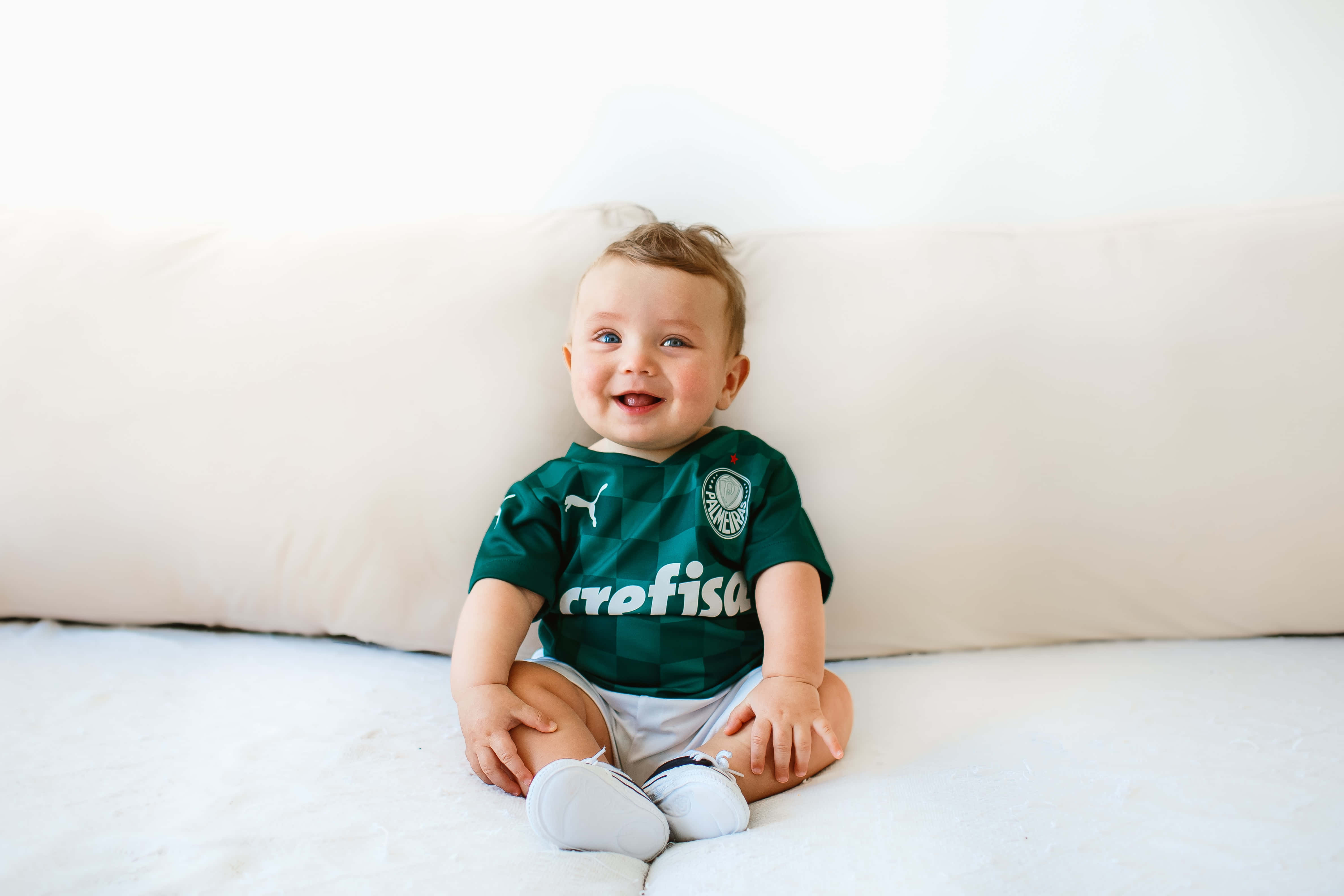 Baby Boy 桌布 壁紙 — 一個快樂的嬰兒穿著綠色足球球衣，坐在白色床上，身後有一個大奶油色枕頭，露出燦爛的笑容。