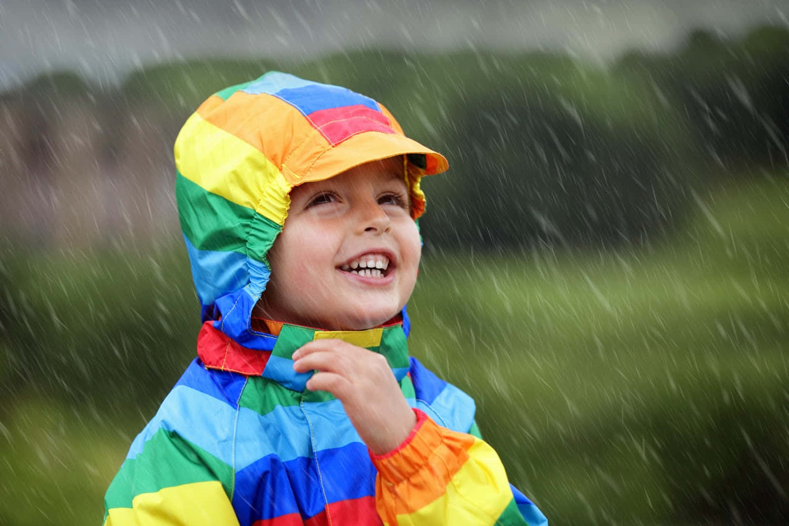 彩色 桌布 壁紙 — 一個快樂的孩子穿著彩虹雨衣，在戶外下雨時開心地大笑。
