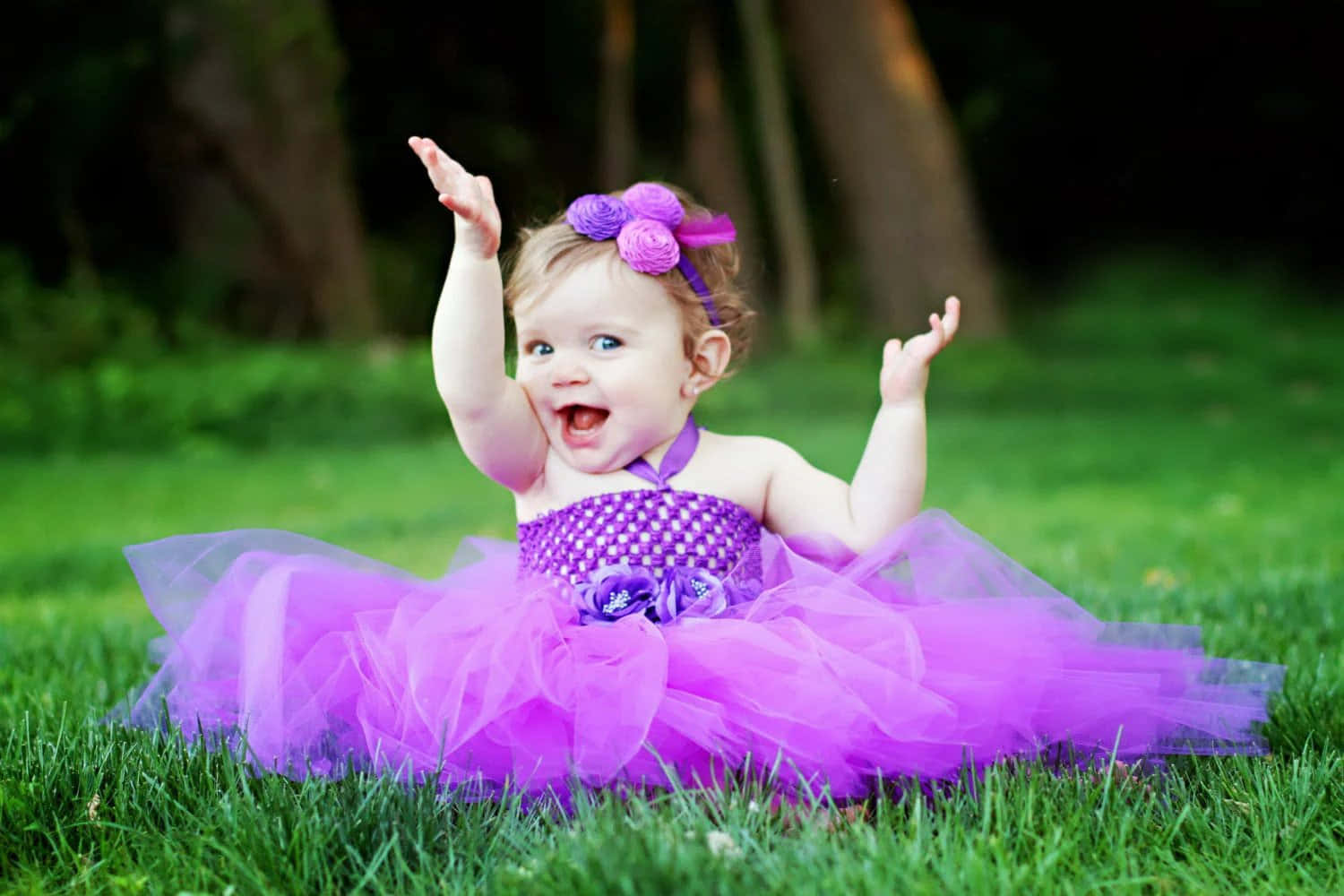 Baby Girl 桌布 壁紙 — 一個快樂的小女孩穿著充滿活力的紫色芭蕾舞裙，坐在茂盛的綠色草地上，高興地舉起雙臂。