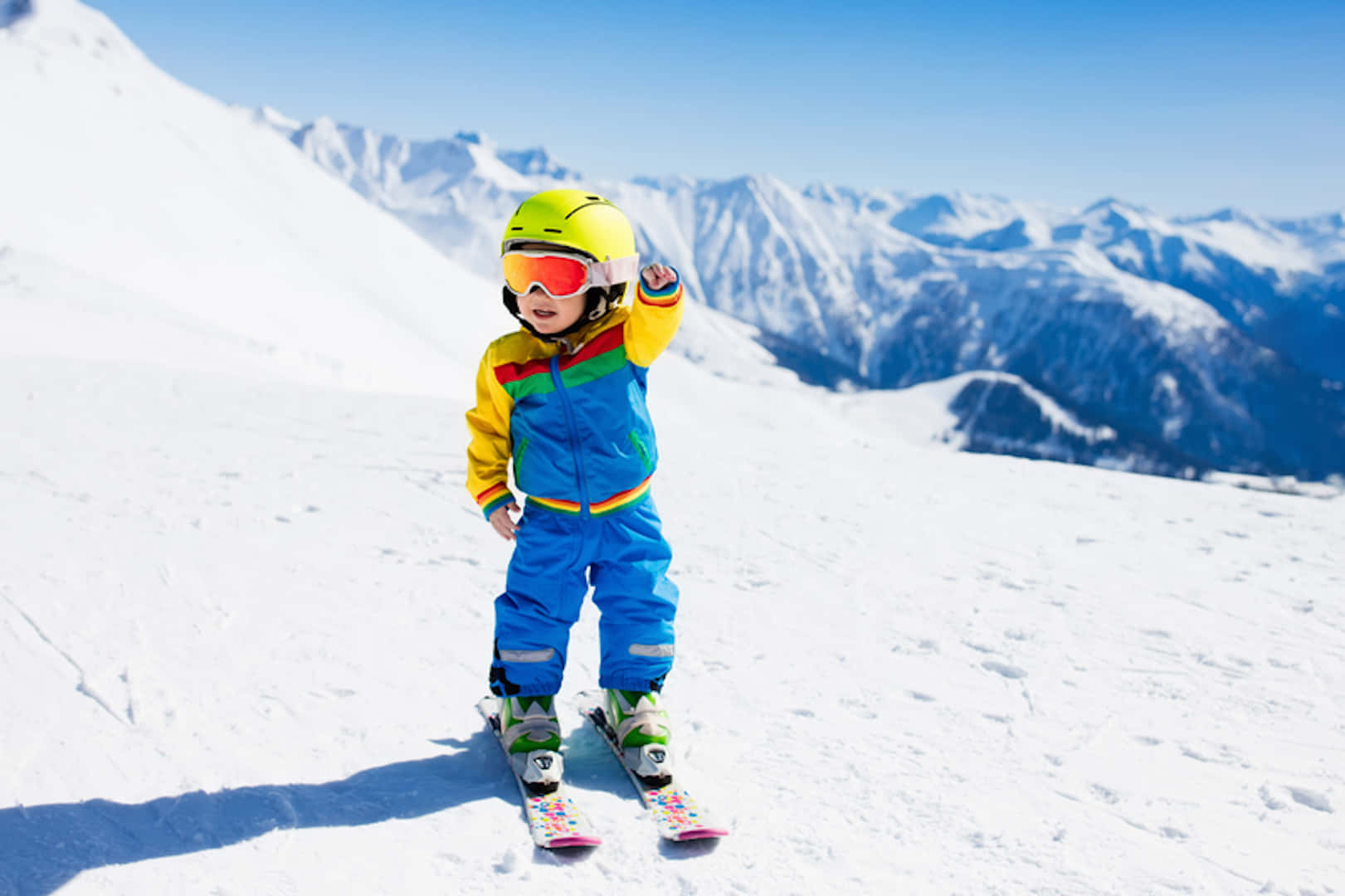 Skiing 桌布 壁紙 — 一個快樂的小孩穿著鮮艷的滑雪裝備，站在陽光明媚的山頂雪地上，背景是白雪覆蓋的山峰。