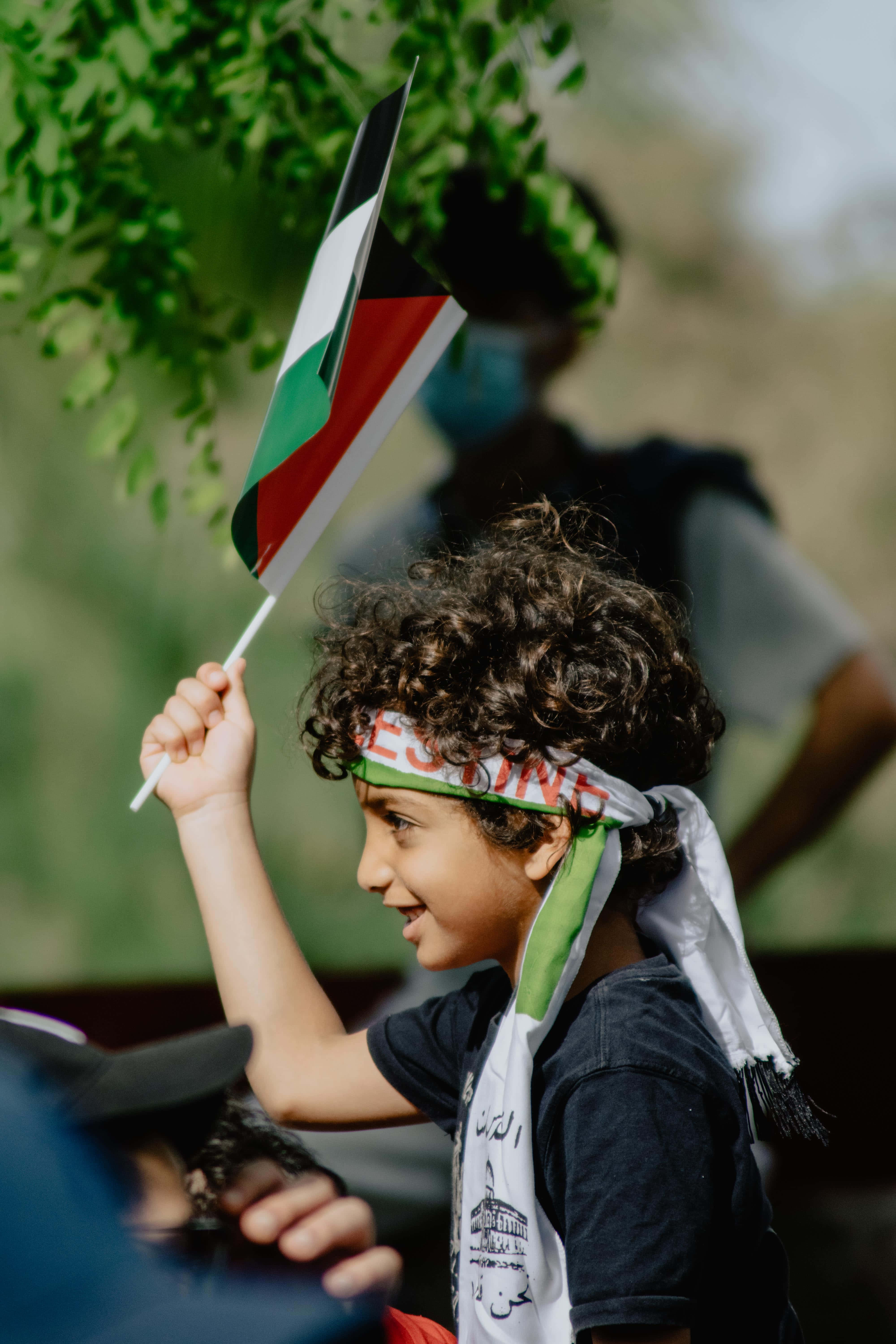 Palestine 桌布 壁紙 — 一個快樂的捲髮小孩在戶外揮舞巴勒斯坦旗幟，戴著相配的頭帶和圍巾。