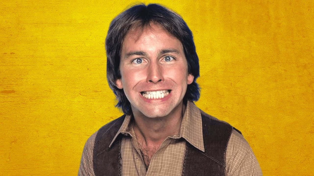 John Ritter 桌布 壁紙 — 一個快樂的男人，擁有明亮的藍色眼睛和燦爛的笑容，穿著棕色背心，內搭格子襯衫，背景是帶有紋理的黃色。
