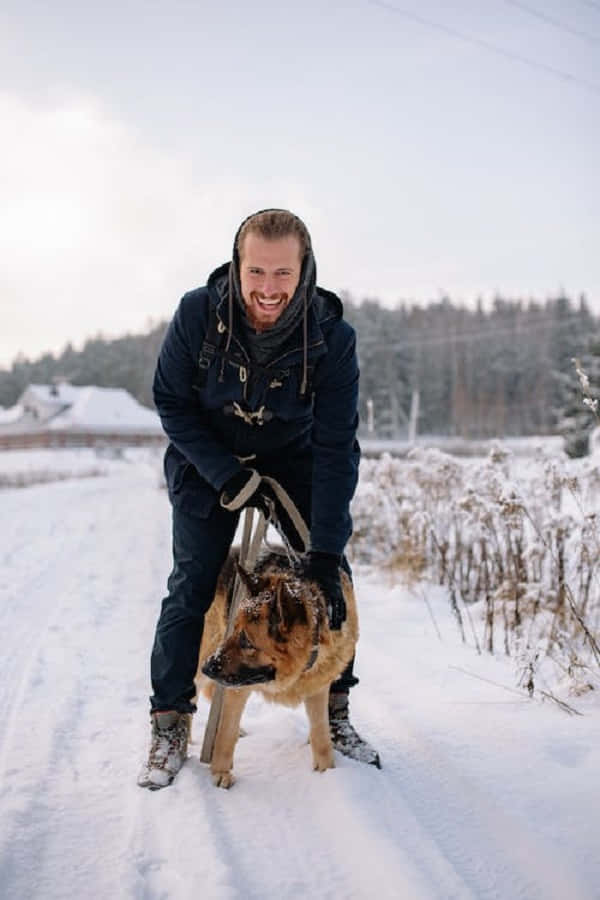 Snowshoes 桌布 壁紙 — 一個快樂的男人穿著冬季裝備，與他的德國牧羊犬站在森林旁邊的雪徑上大笑。