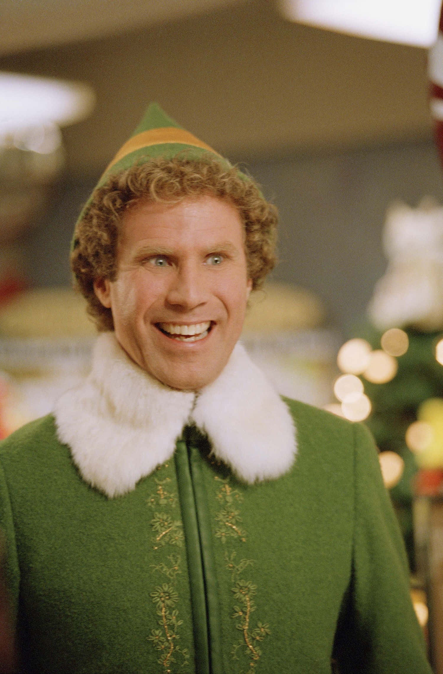 will ferrell 桌布 壁紙 — 一個快樂的男人穿著綠色精靈服裝，配有白色毛領，在室內節日燈光附近燦爛地微笑。