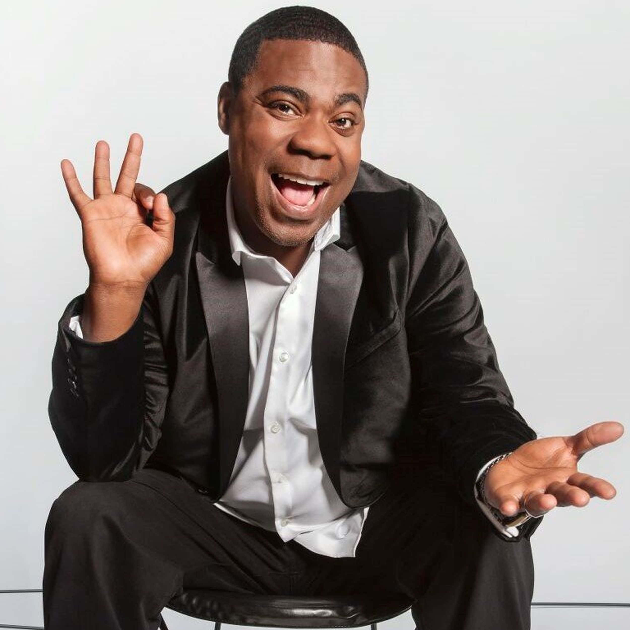 Tracy Morgan 桌布 壁紙 — 一個快樂的男人穿著黑色西裝和白色襯衫坐在凳子上，咧嘴大笑並用手做手勢。