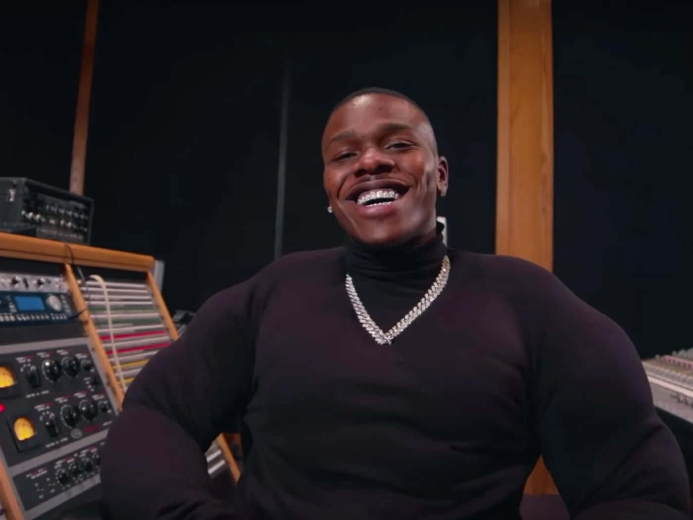Dababy 桌布 壁紙 — 一個快樂的男人穿著黑色高領毛衣和鑽石鏈，在充滿復古音響設備的音樂工作室裡大笑。