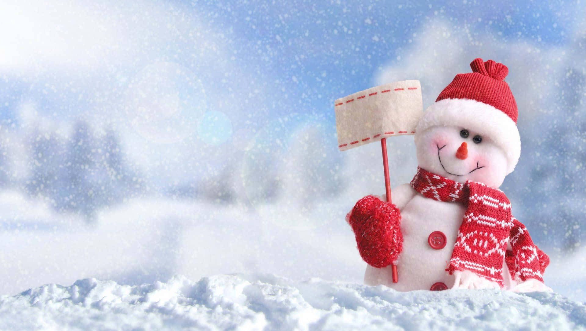 Winter Christmas 桌布 壁紙 — 一個快樂的雪人戴着紅色帽子、圍巾和手套，站在雪景中舉着一個牌子。