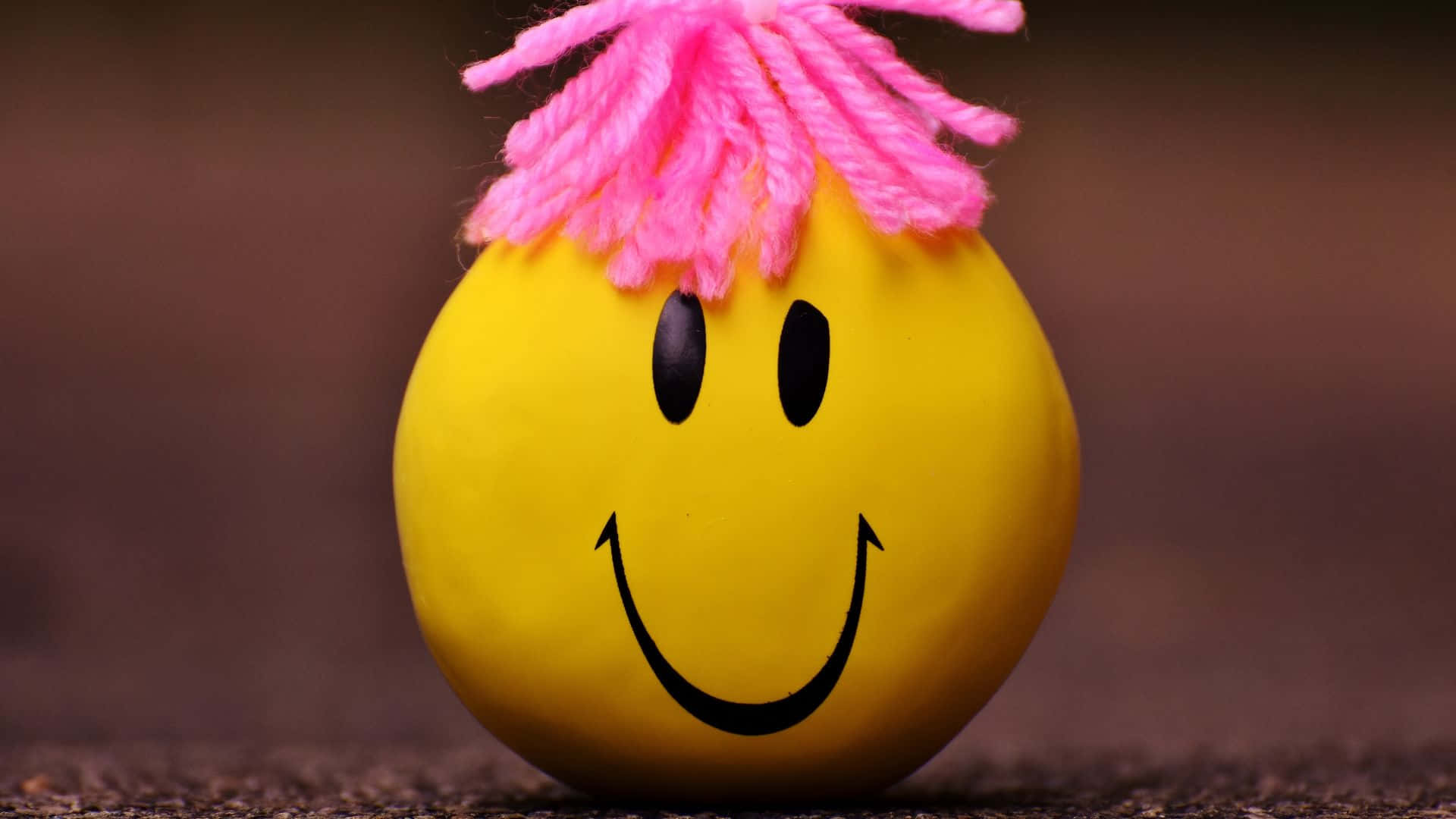 Smiley 桌布 壁紙 — 一個快樂的黃色笑臉球，戴著亮粉紅色紗線假髮，坐在有紋理的地面上，背景是模糊的棕色。