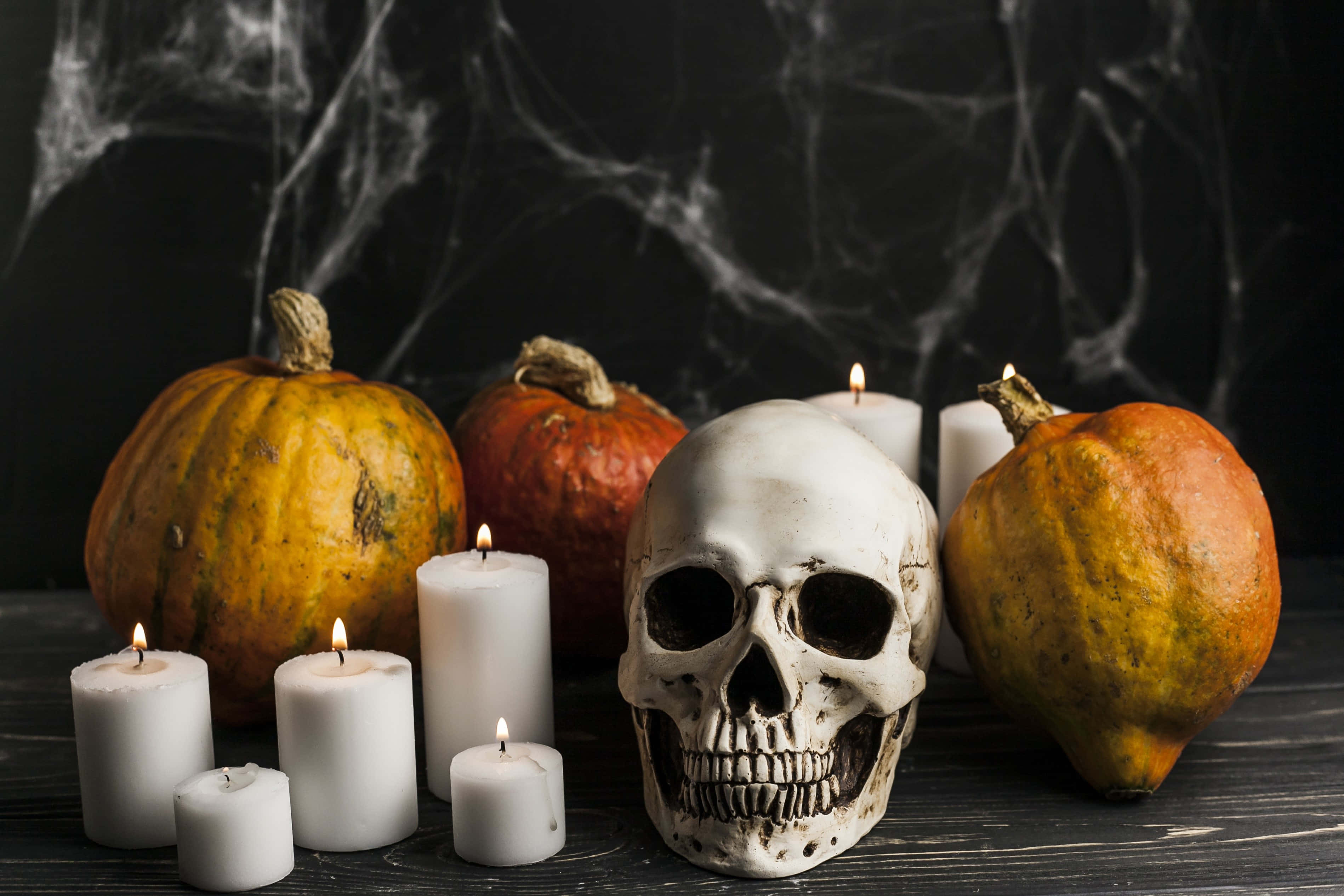 Halloween Props 桌布 壁紙 — 一個恐怖嘅萬聖節靜物畫，有一個人類頭骨被南瓜同閃爍嘅白色蠟燭包圍，背景係黑暗嘅蜘蛛網。