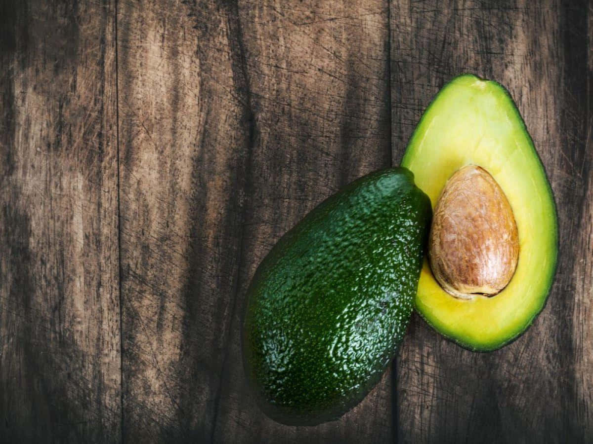 Avocado 桌布 壁紙 — 一個成熟的牛油果切半，露出其奶油狀的綠色果肉和大的棕色果核，放在深色質樸的木質表面上。