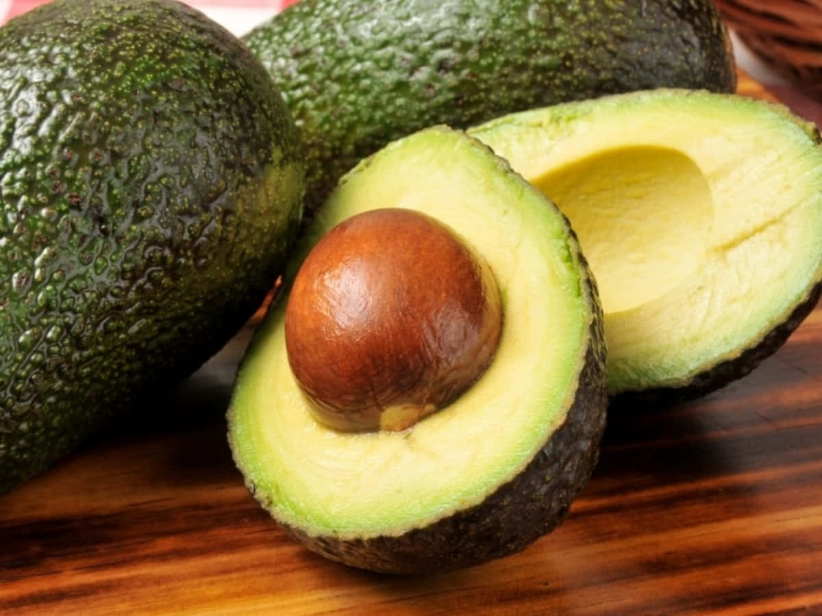 Avocado 桌布 壁紙 — 一個成熟的牛油果切半，露出奶油狀的綠色果肉和一個大的棕色果核，放在木質表面上。