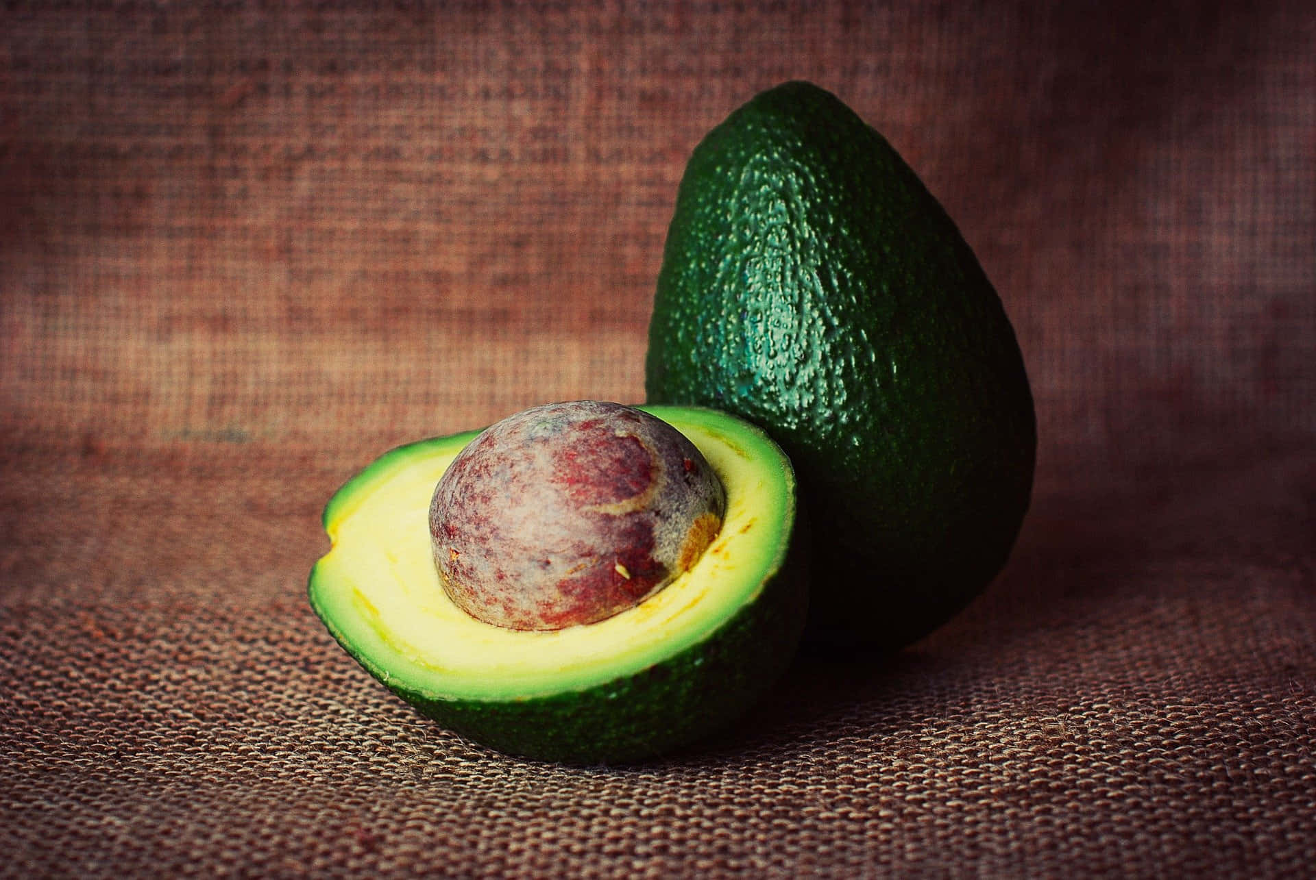 Avocado 桌布 壁紙 — 一個成熟的牛油果切半，露出奶油黃色的果肉和大的棕色果核，放在有紋理的棕色粗麻布表面上。