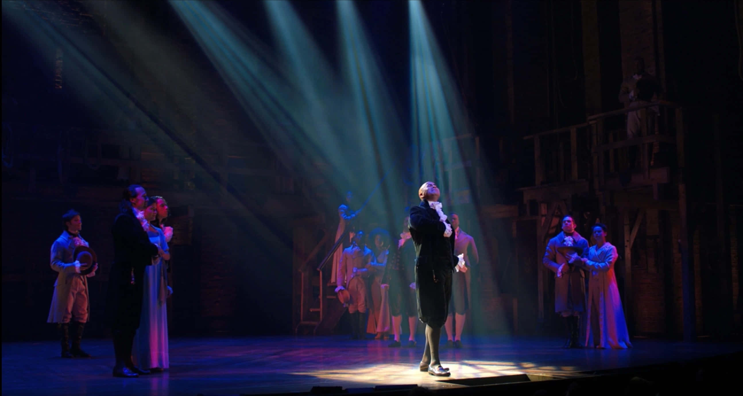 Musical 桌布 壁紙 — 一個戲劇性的劇院舞台，沐浴在聚光燈光束中，演員穿著古裝在昏暗的環境燈光下表演。