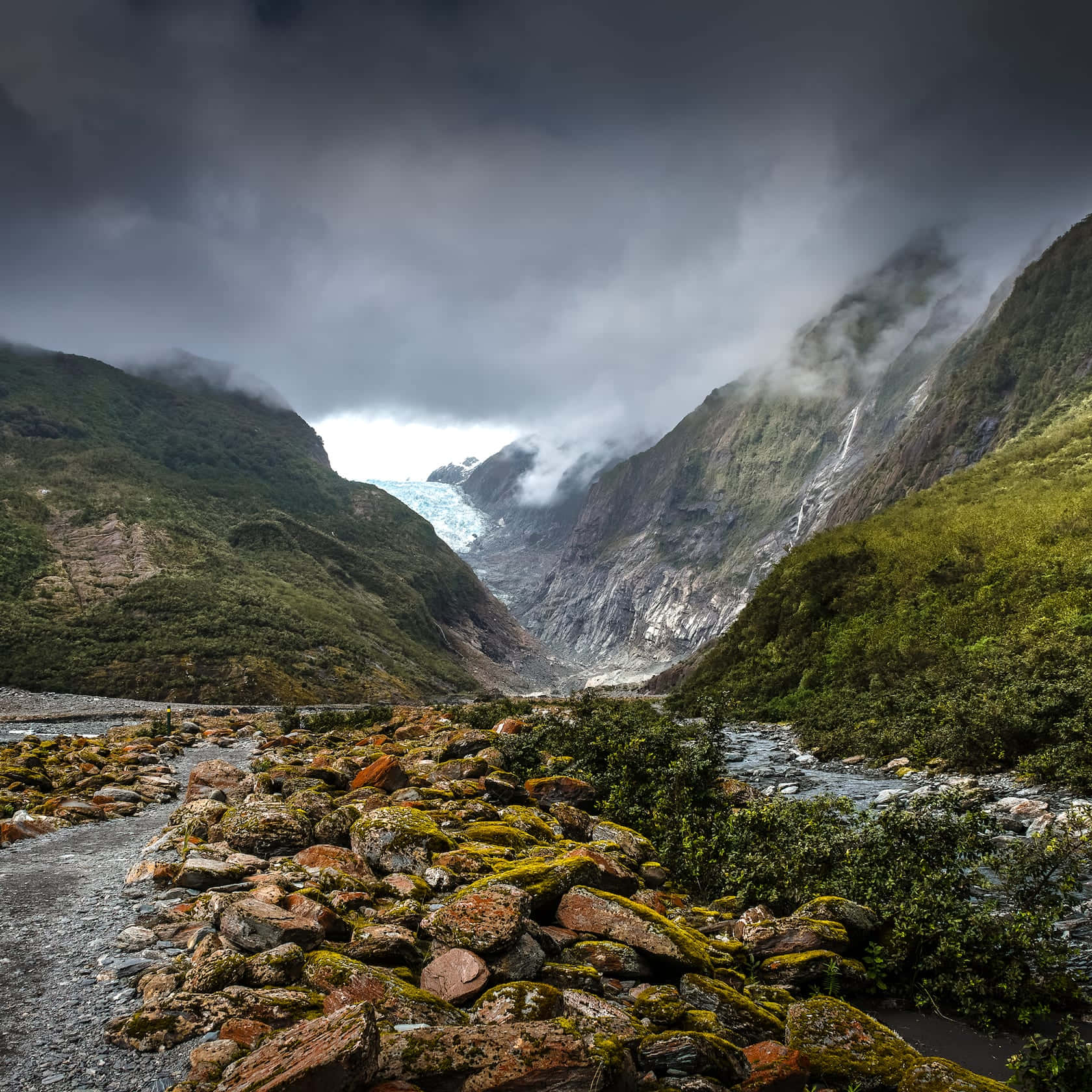 New Zealand 桌布 壁紙 — 一個戲劇性的山谷，佈滿苔蘚的岩石和一條河流，通向暴風雨天空下的遠處冰川。