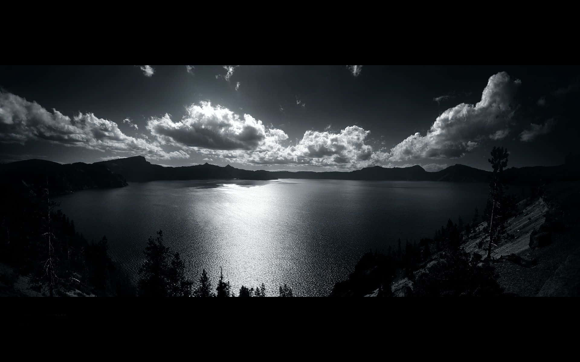 Black And White Landscape 桌布 壁紙 — 一個戲劇性的黑白全景，展示廣闊湖泊透過暴風雲反射陽光，帶有剪影樹木和遠山。