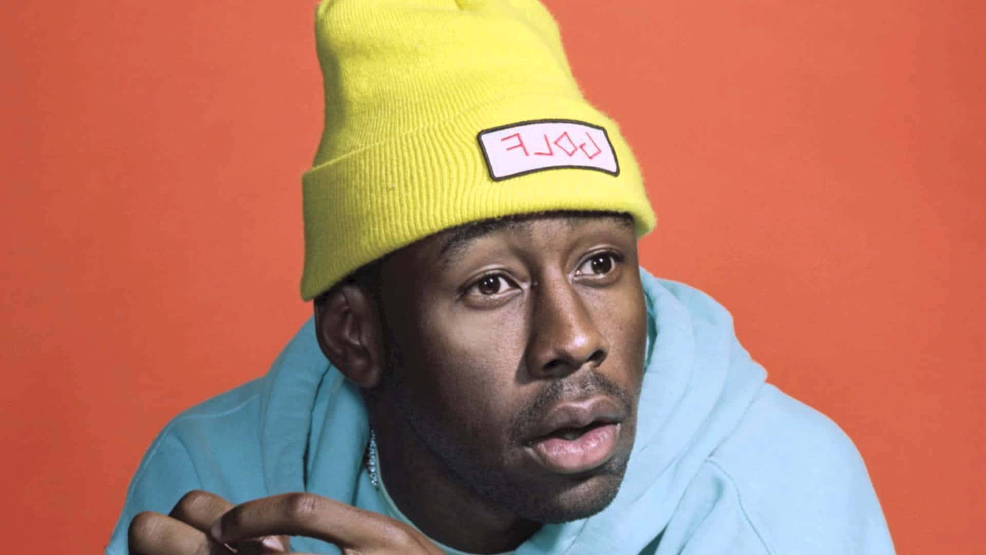 tyler the creator 桌布 壁紙 — 一個戴住黃色冷帽同淺藍色連帽衫嘅男人，喺純橙色背景前專注咁望住。